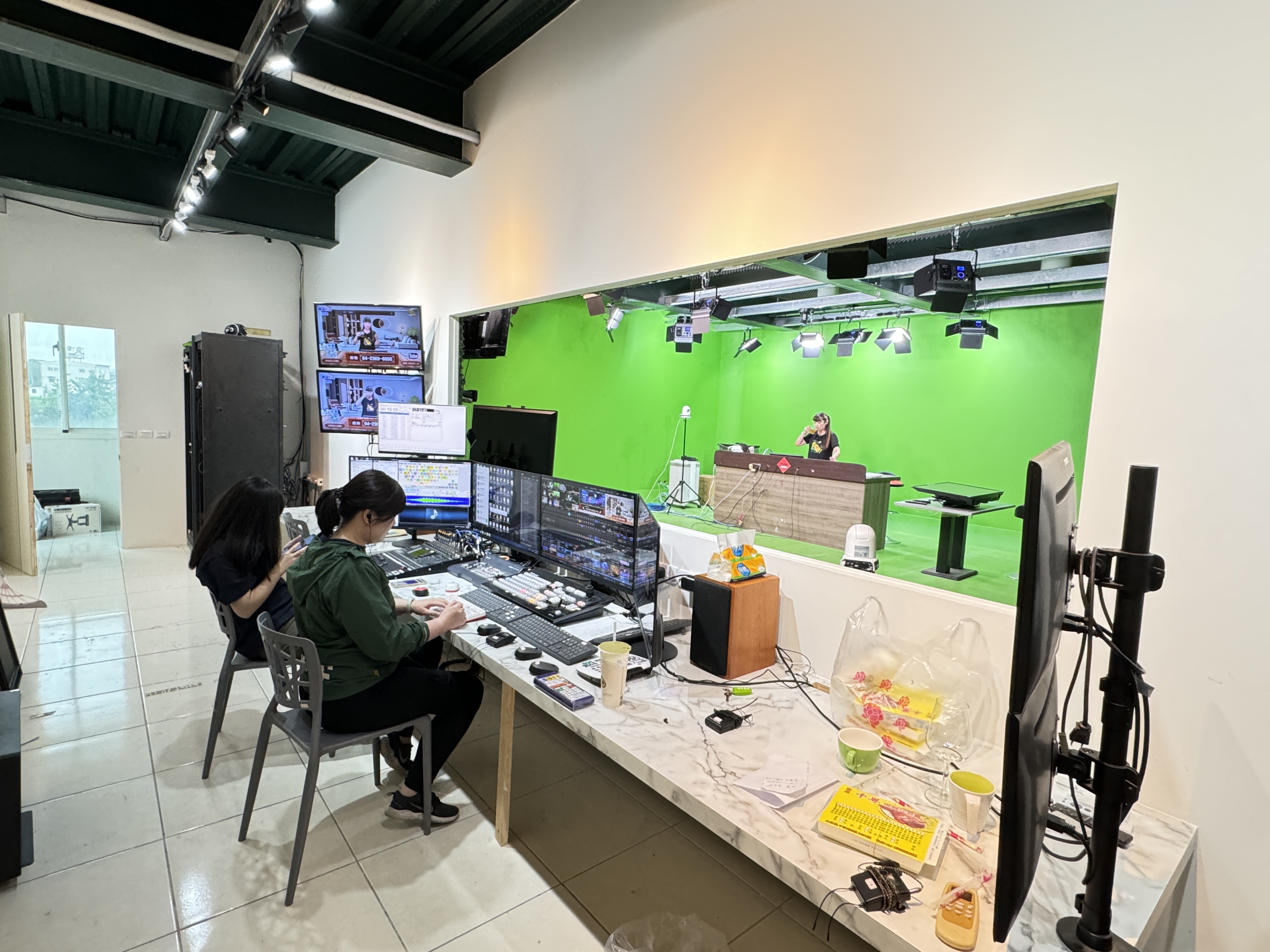 工作人員坐在多螢幕導播桌前，透過前方大型開口可看見綠幕棚、棚燈及棚內人員。